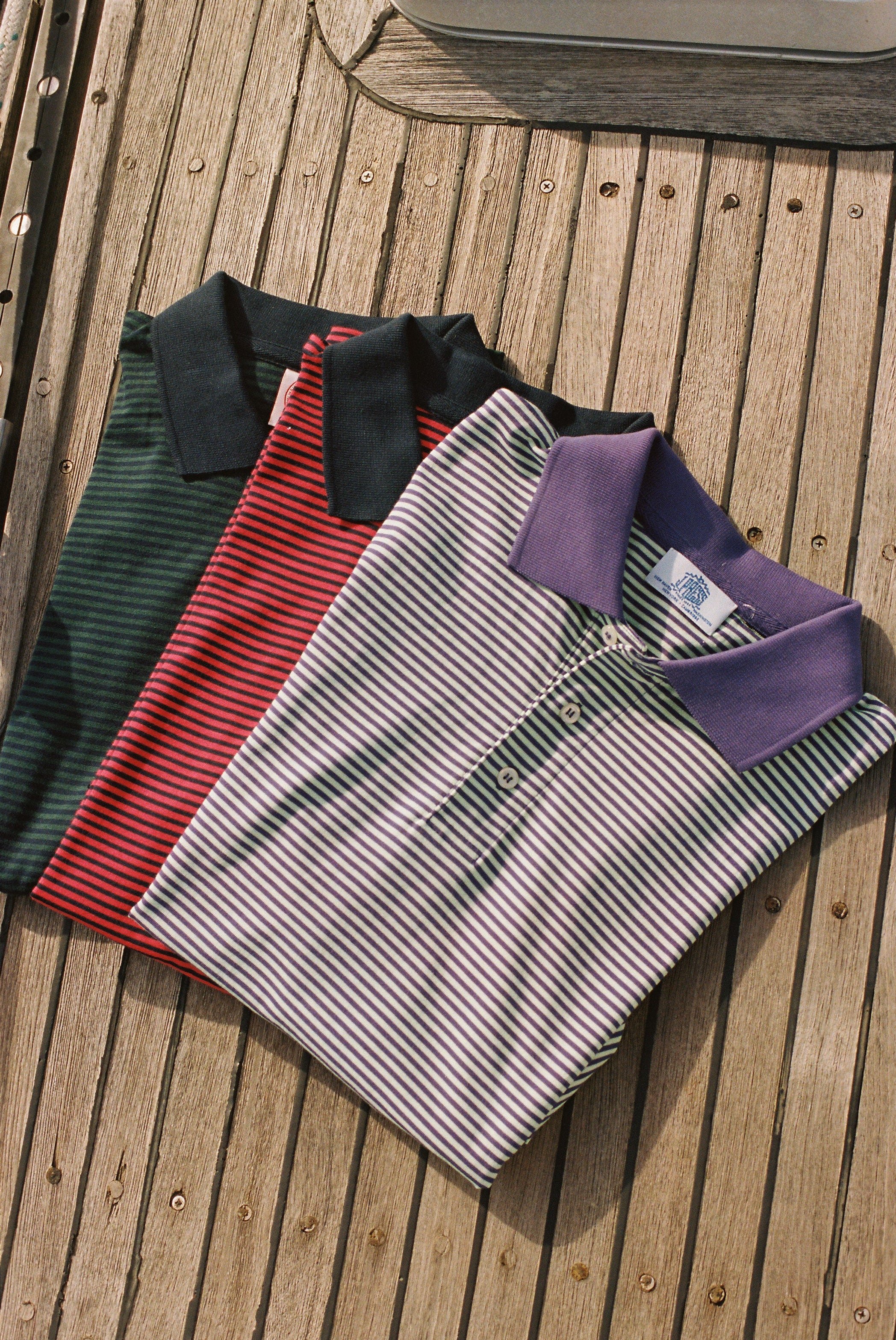 Three folded striped polo shirts are stacked diagonally on a wooden slatted surface. The top shirt is white with thin green stripes and a purple collar; the middle is red with thin green stripes and a dark green collar; the bottom is dark green with thin red stripes and a dark green collar.