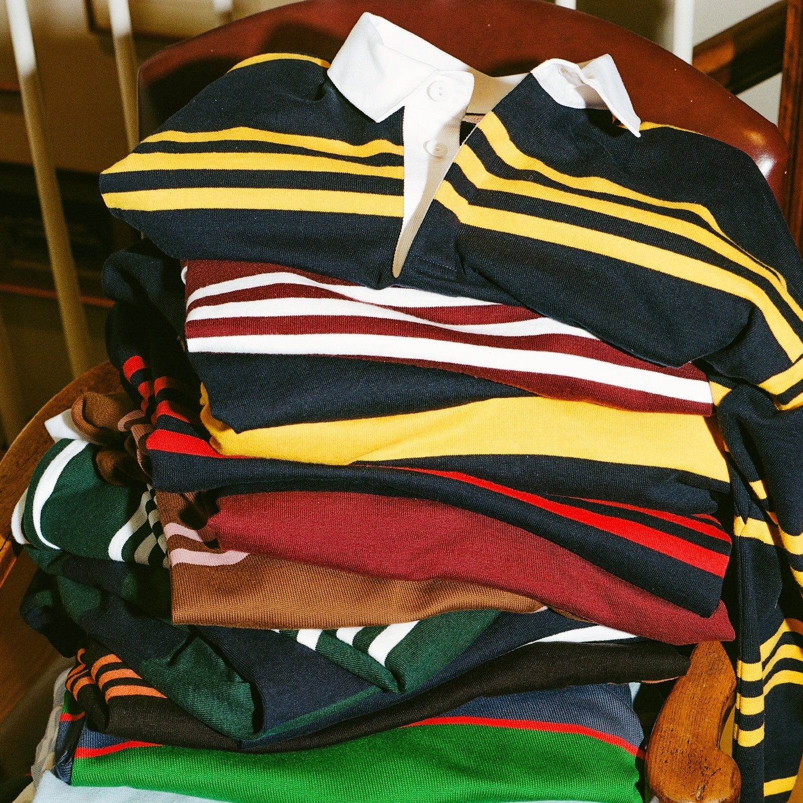 A stack of folded striped polo shirts in various colors, including navy with yellow and white stripes, and solid red, green, and blue shirts, arranged on a wooden chair.