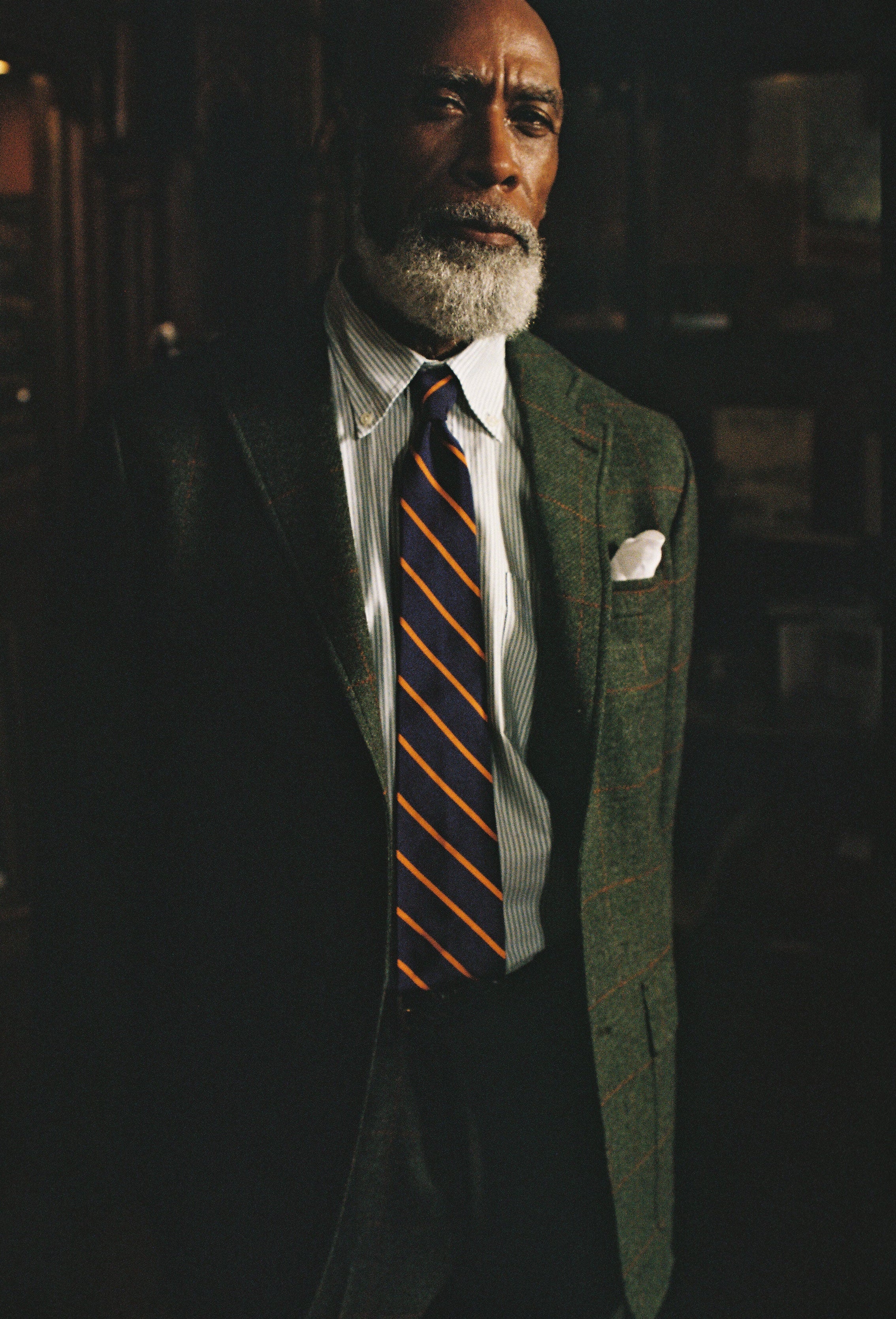A portrait of a man with a white beard wearing a dark green patterned suit jacket, a white shirt, and a navy tie with orange diagonal stripes, set against a dark, moody background.