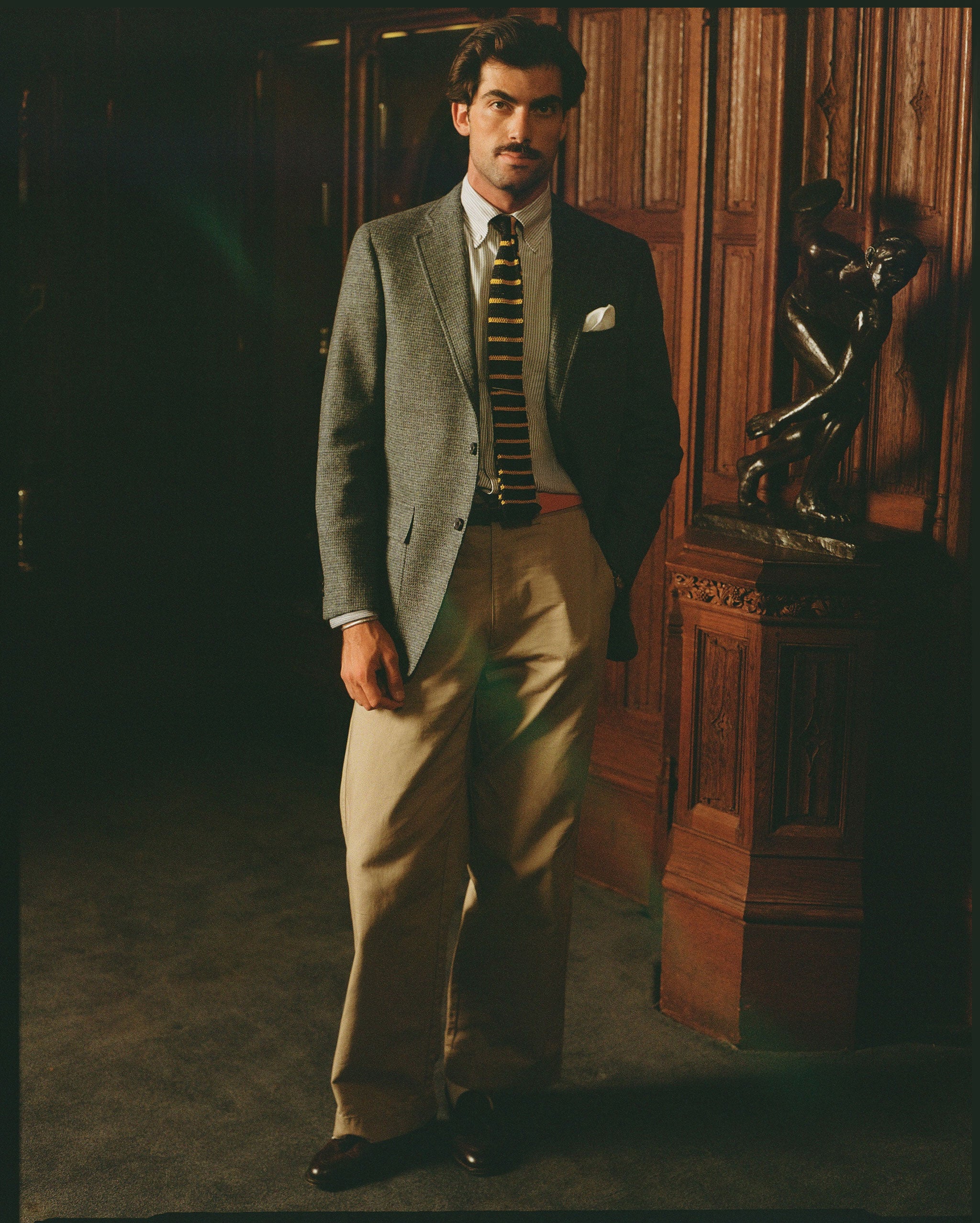 A man stands in a dimly lit interior with wood-paneled walls and a pedestal, wearing a gray blazer, beige trousers, a striped tie, and dark loafers.